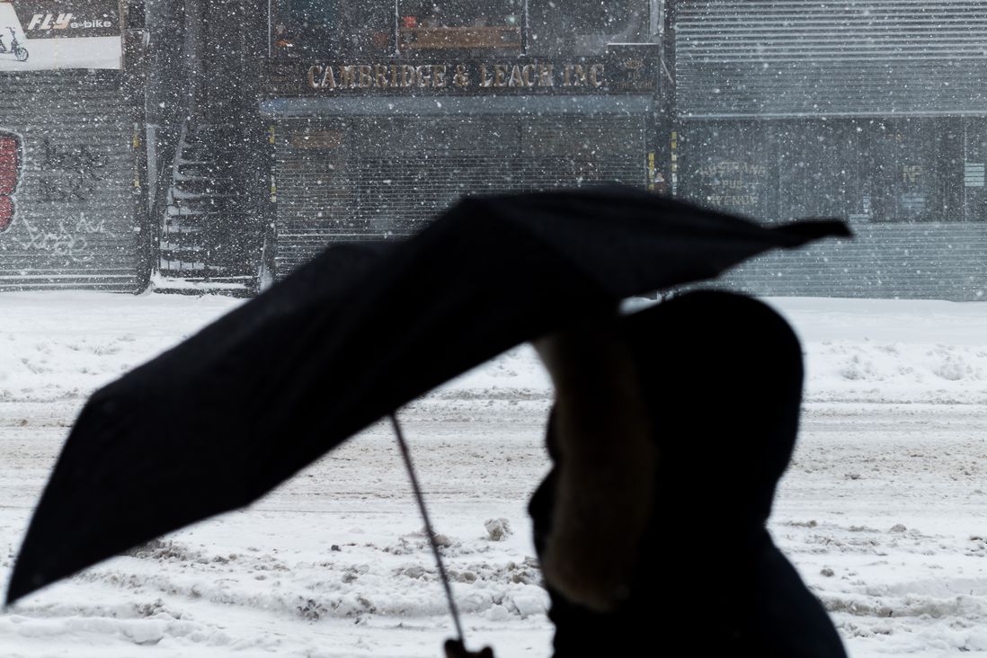 A person holding an open umbrella in the foreground with shuttered stores and snow falling in the background
